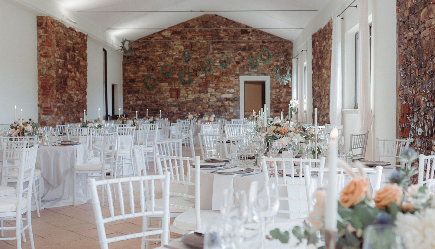 Sala allestita per ricevimento cerimonia all'interno della tenuta: tavoli e sedie bianchi e centrotavola floreali
